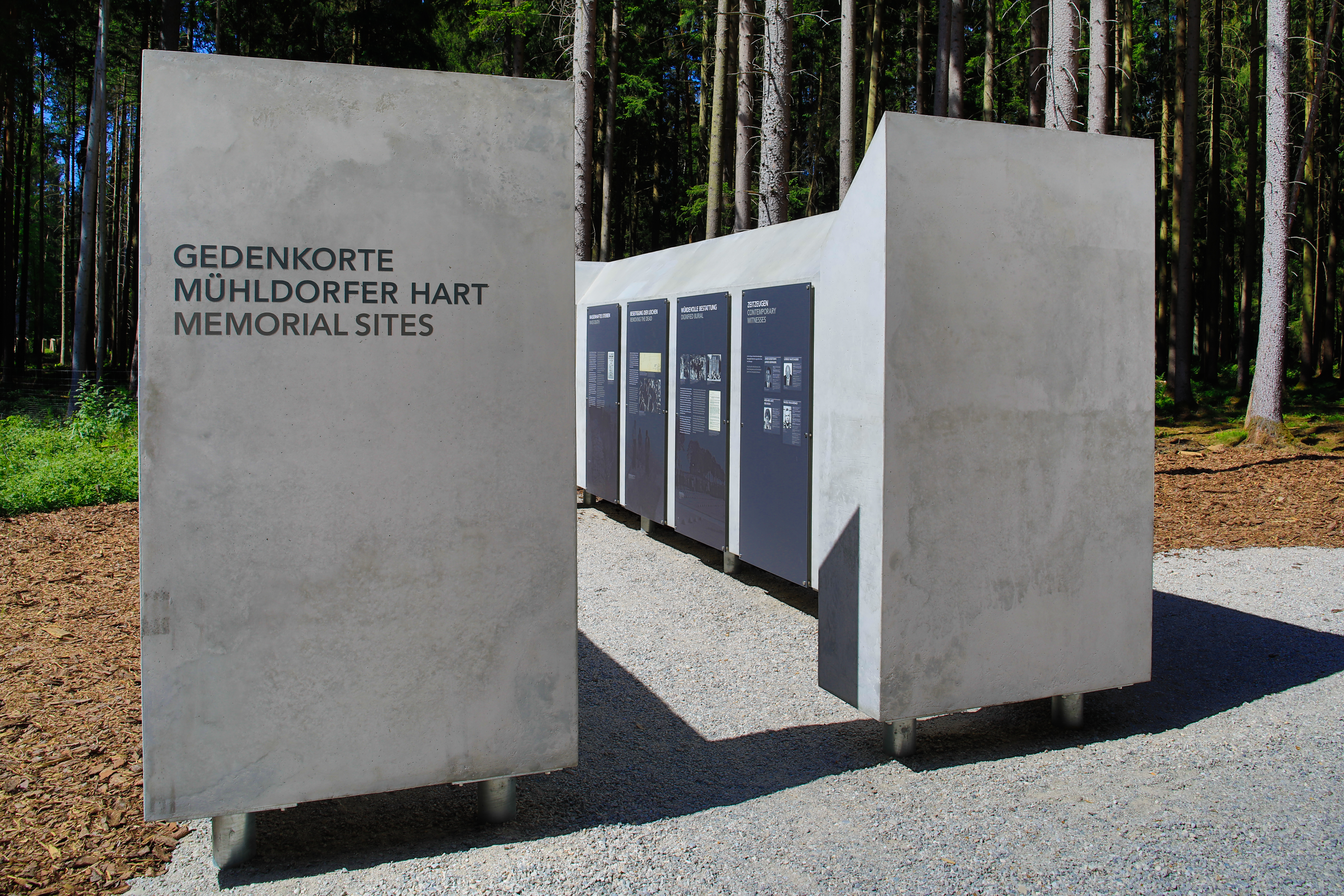 Foto: Stiftung Bayerische Gedenkstätten / Heiner Heine, Burgkirchen Fotografie von Gedenktafeln aus Beton im Wald, die an die Mühldorfer Hart Memorial Sites erinnern. Tafeln zeigen mehrsprachige Inschriften und historische Informationen, angeordnet in einer offenen, begehbaren Struktur.